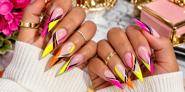 Long neon stiletto acrylic nails featuring vibrant pink, yellow, orange, black, and white geometric line accents on a glossy nude base, styled with gold rings.