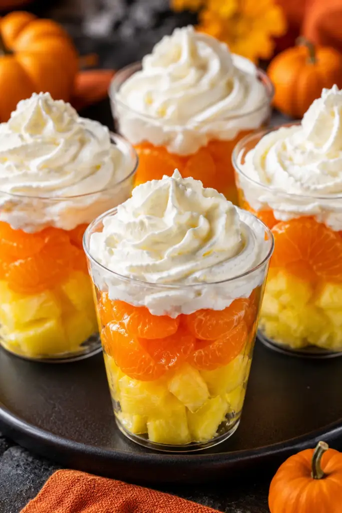 Bright candy corn fruit parfaits layered with golden pineapple, orange mandarin pieces, and fluffy whipped cream in clear cups for a festive Halloween party.